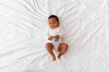 Charming Little Black Baby With Rattle Toy In Hands Relaxing In Bed