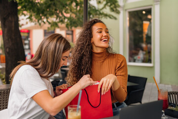 Two happy girls are sitting in the cafe and checking the red shopping bag to see what they have bought.