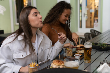 Two girls are eating delicious burgers with lots of toppings and french fries, one of them is really delighted by its taste. They are drinking beer, too.