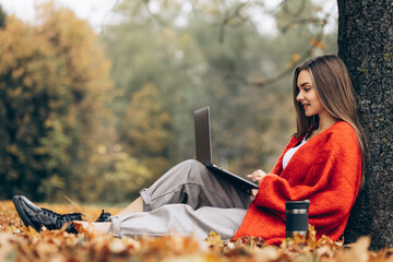 Woman sitting under the tree in park working on laptop and drinking coffee