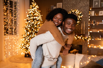 Happy millennial african american husband hold wife on back, have fun in cozy living room interior