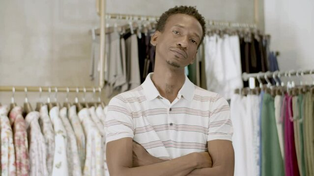 Bearded black man standing with folded arms in clothing store, looking at camera and smiling. Racks of colorful clothes in background. Guy having BLK PWR tatoo on neck. Shopping, lifestyle concept.