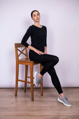 Fototapeta na wymiar full length woman poses on a high wooden chair in black tight fitting clothes and sneakers in a studio on a white background.