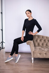 full length woman poses on a sofa in black tight fitting clothes and sneakers in a studio on a white background.