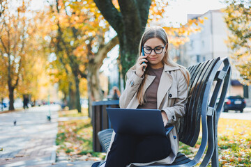 A smiling woman works with a laptop on a bench in the Autumn Park. She has a great smile, long hair and big blue eyes. Portrait of a modern working woman. Yellow park background