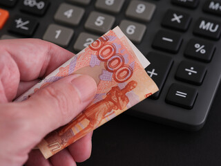 A man holding a five thousand ruble banknote in his hand with a calculator in the background