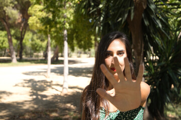 Young woman, brunette, slender, dressed in green t-shirt and jeans, with her hand covering the camera. Concept stop, no, denial, refusal. Selective focus on face.