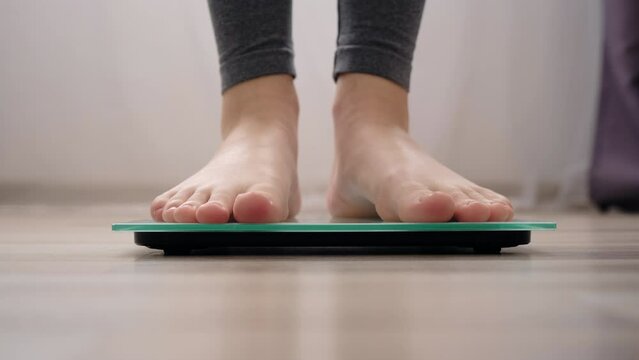 Selective focus of female feet weighing on scales in living room at home. Female dieting checking and weight loss. Barefoot measuring body fat overweight. Control, wellness and diet concept