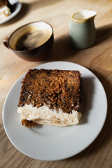 Shallow focus on the icing of a slice of carrot cake. It is on a wooden table lit by window light which is creating with interesting shadows. There is a cup of black coffee and milk jug too.