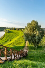 A wooden gangway of an old fortress on the river bank among lush trees and rural houses. Scenic landscape with green grass and blue sky in summer evening