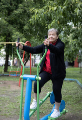 Smiling elderly woman is doing exercises on simulator, trainer ellipse outdoors in the yard. Active life of pensioners. Adaptation of pensioners in the modern world. Prevention of mental illness.