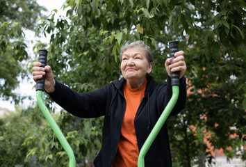Smiling elderly woman is doing exercises on simulator, trainer ellipse outdoors in the yard. Active life of pensioners. Adaptation of pensioners in the modern world. Prevention of mental illness.