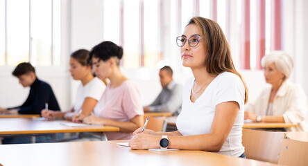 Focused young attractive Hispanic woman listening to lecture and taking notes in classroom with...