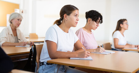 Young woman sitting at desk in classroom and writing while listening lecture on foreign language...