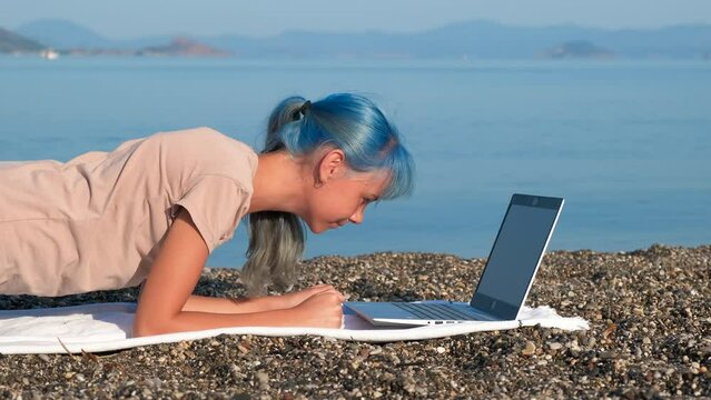 Girl stand in the plank on the beach. A happy teen stay in plank by her laptop with sport exercises on the pebbles luxury beach by the blue ocean.