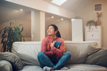 Asian woman siting on the couch at home and holding a book in her arms