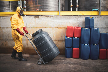 Chemical worker in yellow hazmat suit and respiratory mask moving acid barrels to warehouse...
