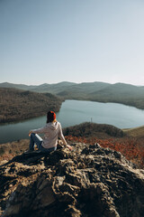 young woman in sportswear and hat is sitting on top of mountain and looking at autumn landscape and lake. independent tourism. unpopular vacation spots. vertically, selective focus
