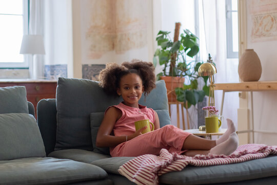 Portrait Of African American Girl In Pink Overall. Cute Child With Curly Hair Drinking Hot Cocoa Milk Or Tea From Yellow Mug. Portrait, Childhood Concept