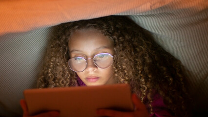 Close-up of little fair-haired girl holding tablet. Female child in glasses sitting quietly in bed...