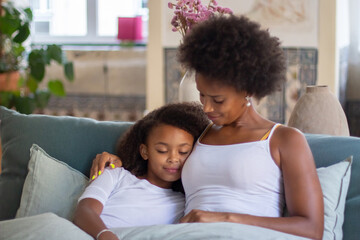 Mother and daughter with curly hair cuddling in bed. African American woman and little girl sitting, leaning on shoulder, eyes closed. Family, parenthood, home concept