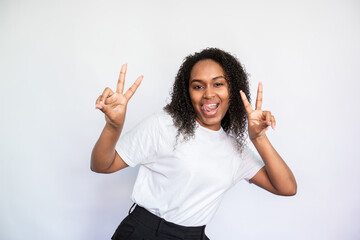 Portrait of cheerful young woman making victory gesture and sticking out tongue. African American lady wearing white T-shirt looking at camera with joyful expression. Fun and happiness concept
