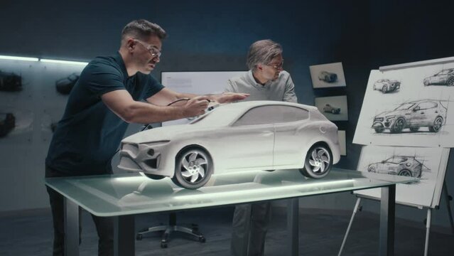 Senior automotive design engineers discuss the design of the car looking at the pencil sketches while working on the sculpture model of the car. Using a rotary tool for final touch ups.