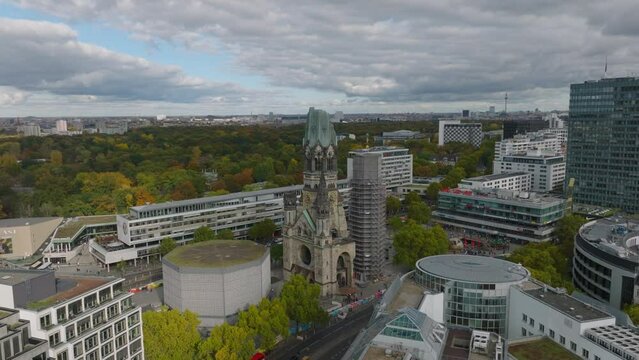 Aerial ascending footage of famous historic landmark. Historic church between modern development and autumn colour trees in park in background. Charlottenburg neighbourhood, Berlin, Germany