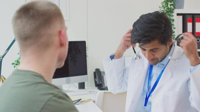 Male doctor or GP wearing white coat examining young man listening to heartbeat with stethoscope- shot in slow motion