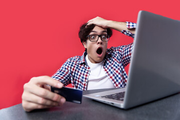 Shocked man gasping in surprise with his credit card balance, looking at his finances on his laptop