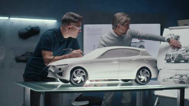 Two experienced automotive engineers discuss the car design looking at the sketches on the board. Making corrections in the prototype rake sculpture using a rotary tool with spinning tip.