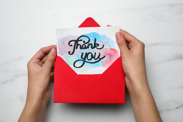 Woman holding envelope and card with phrase Thank you at white marble table, top view