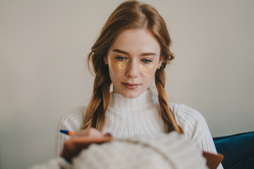 Beautiful young woman with under eye patches holding pen with notebook and sitting on sofa at home office. Education concept. Relaxing time.