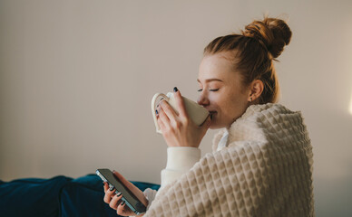 Young woman sitting on the sofa with plaid, drinking coffee, relaxing in her living room. Happy...