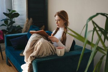 Beautiful woman with cosmetic eye patches sitting on the sofa in cozy clothes and writing in...