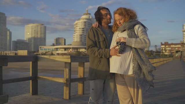 a couple in love stands at the end of a pier near the beach and kisses while the man gently rubs his pregnant wife's belly.