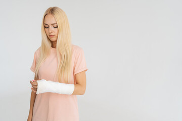 Studio portrait of unhappy injured blonde young woman with broken arm wrapped in plaster bandage...