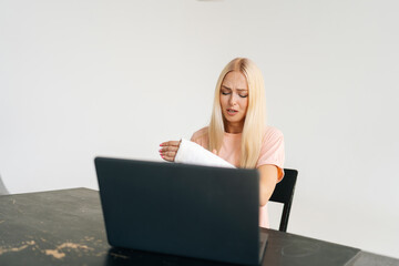 Studio portrait of suffering from pain young woman with broken arm wrapped in plaster bandage having video call, online consultation on laptop sitting at black table, on white isolated background.