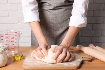 Obraz na płótnie Canvas Woman kneading dough at wooden table near white brick wall, closeup