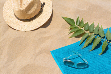 Soft blue beach towel, sunglasses, straw hat and green leaves on sand, above view