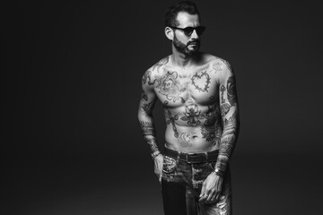 Fototapeta na wymiar Portrait of handsome confident stylish hipster lambersexual model. Sexy modern man. Naked torso with tattoos.Fashion male posing in studio on dark background in sunglasses