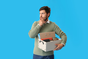 Sad fired young man holding box with personal stuff on blue background