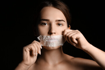 Young woman with taped mouth on dark background. Censorship concept