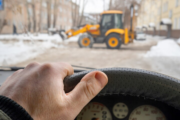 view of the car driver hand on the steering wheel against the background of a tractor in front of the car, shoveling a snow blockage and blocking the road, creating a traffic jam on the roadway