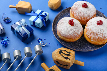 Plate with tasty donuts, dreidels and gifts for Hanukkah celebration on blue background, closeup