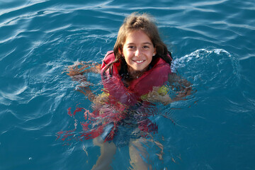 A child in a life jacket swims in the sea. Girl with long dark hair in blue water.