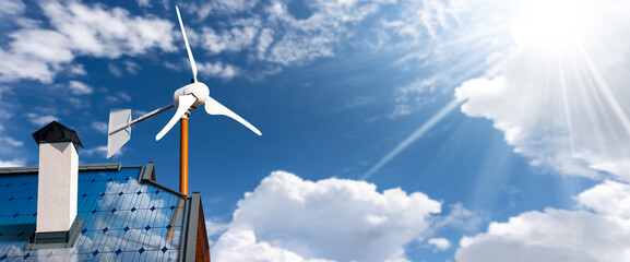 Solar panels and a small wind turbine on the top of a roof of a house, against a blue sky with...
