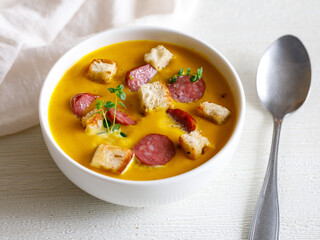 Homemade spicy Pumpkin cream soup, served with smoked mini sausage and bread on a white wooden table with spoon. Top view close up