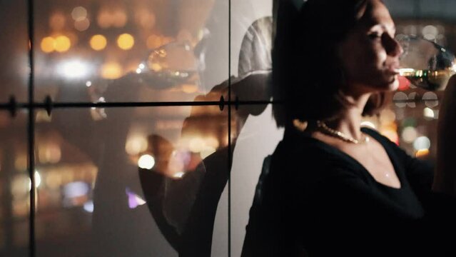 Elegant brunette woman drinking white wine glass at rooftop bar terrace looking at city lights. Sexy female hanging out. Big city night concept.