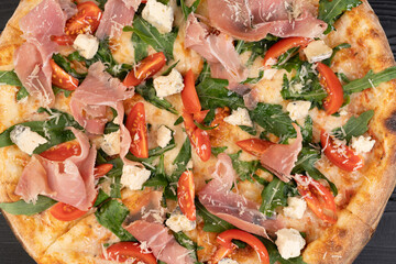 Close up of pizza with prosciutto, tomatoes, arugula and parmesan on wooden background, top view.